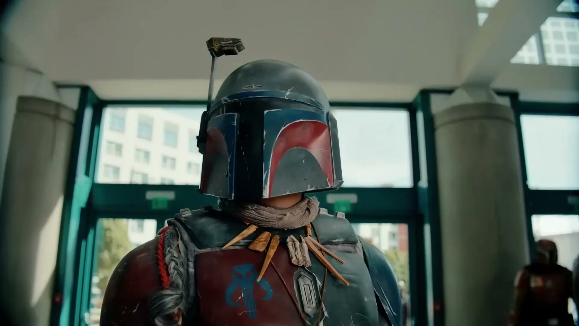 A person in a detailed Mandalorian costume with a helmet and tribal necklace stands in a bright, modern building entrance.