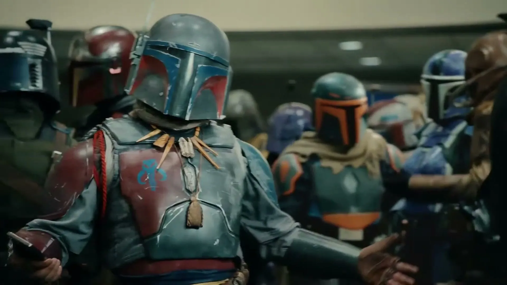 A person dressed as a Mandalorian in full armor looks at their phone, then looks up and gestures, surrounded by other Mandalorians at a convention.