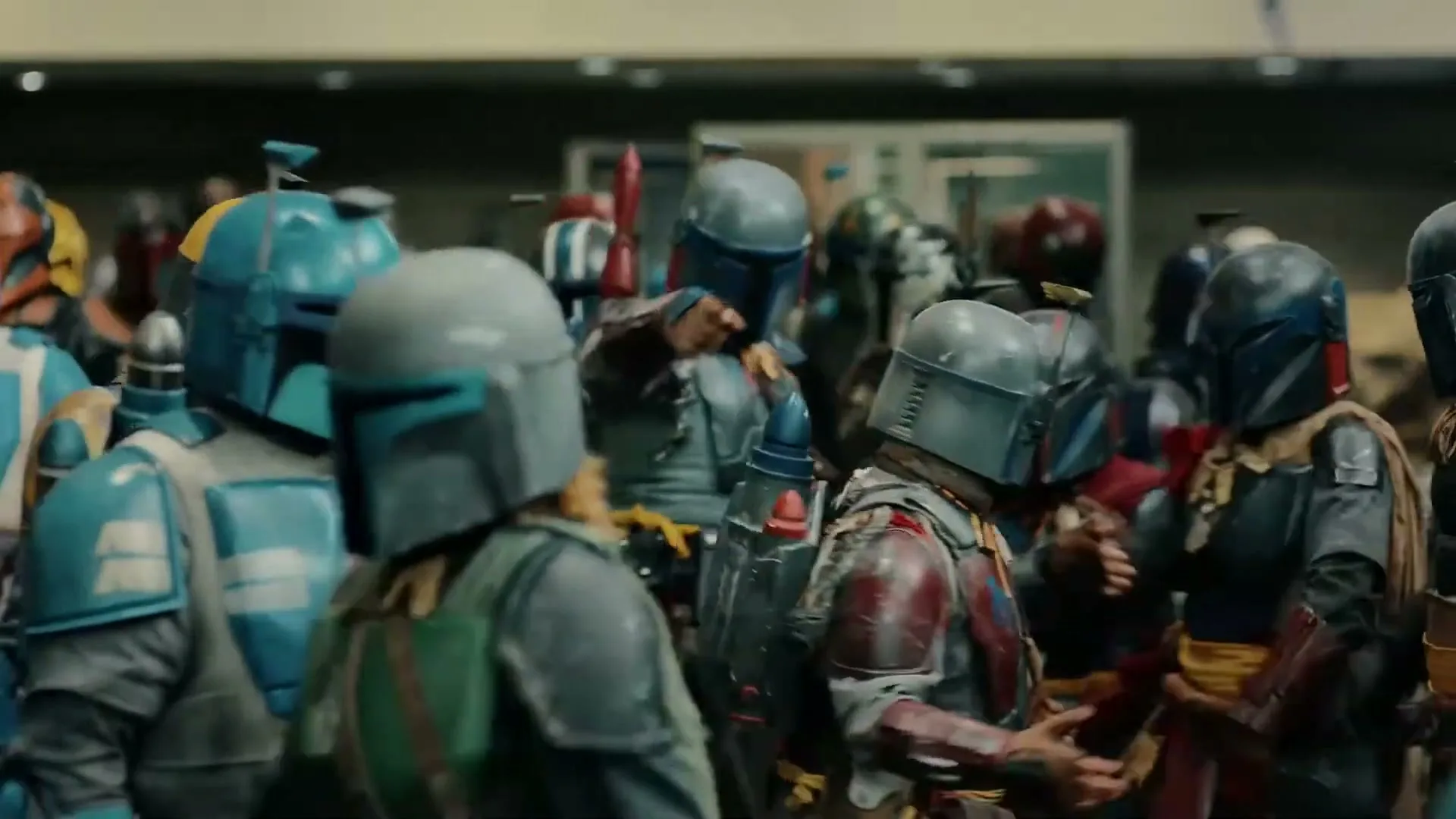 A large group of people dressed in colorful Mandalorian armor walk through a convention hall, some turning their heads as if looking for someone.