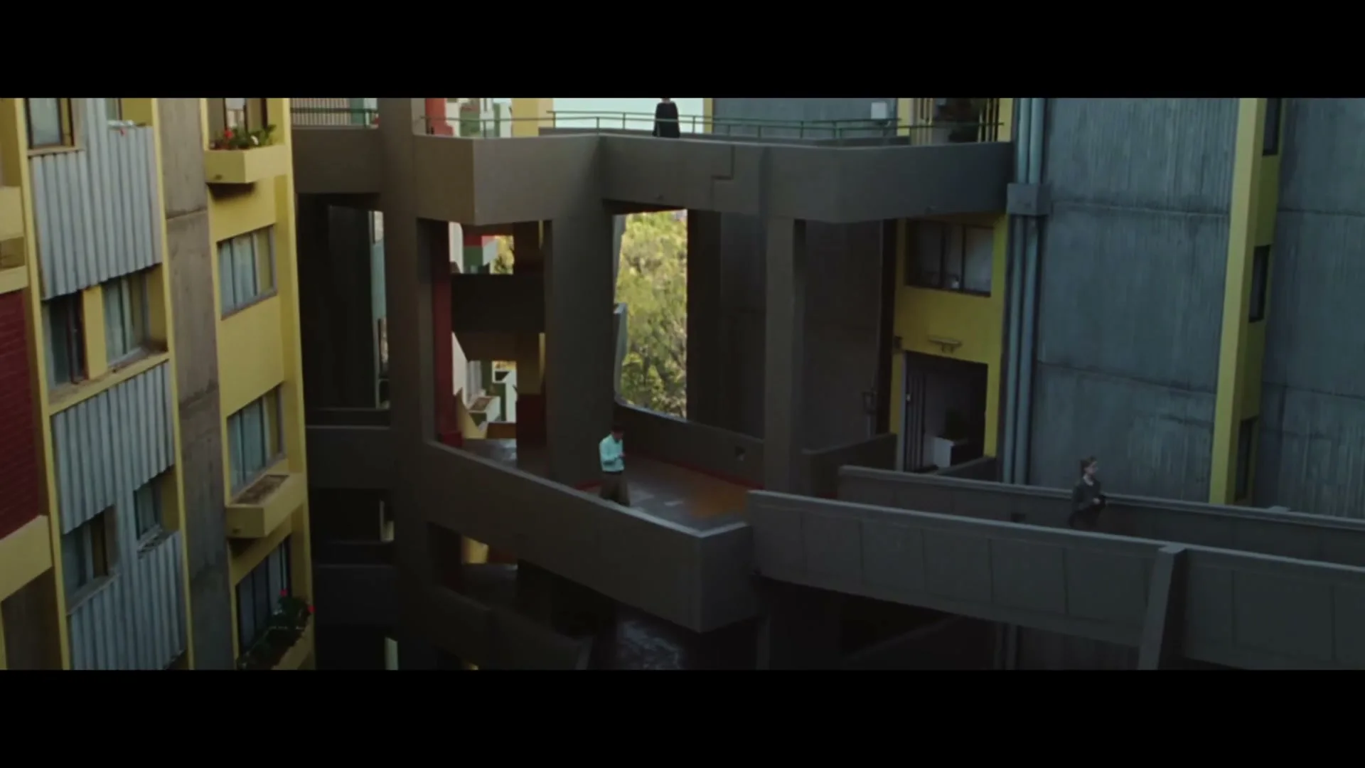 A wide shot of a brutalist apartment complex with yellow and grey buildings, connected by walkways, as people walk across them.