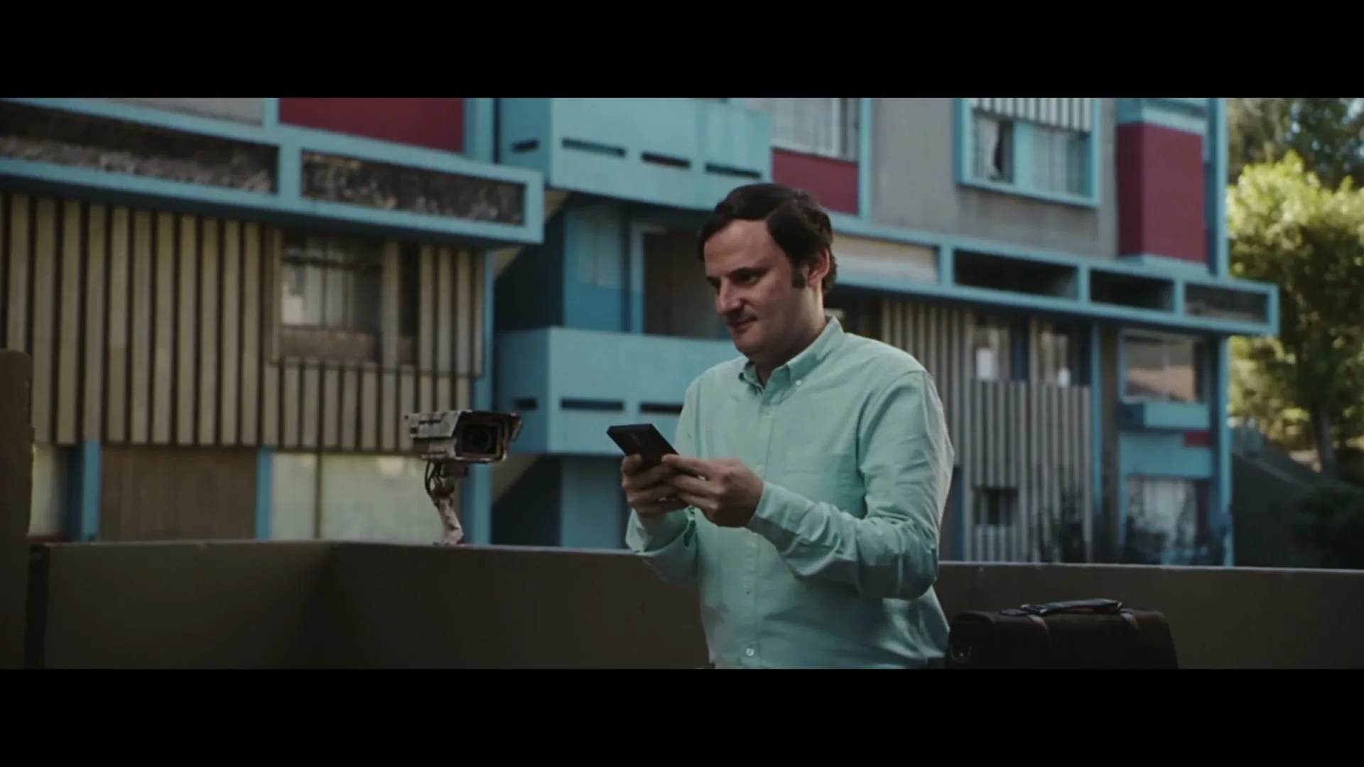 A man in a light green shirt stands on a balcony, looking at his phone, as a security camera robotically turns to face him.