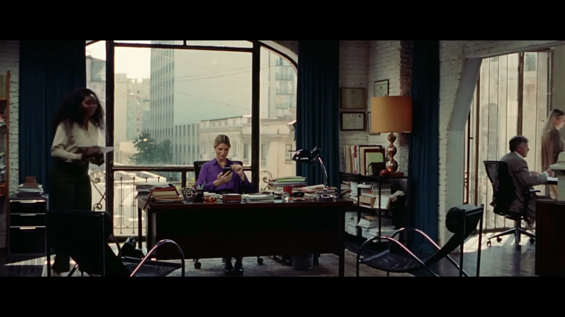 A woman in a purple shirt sits at a desk, looking at her phone, while another woman walks past her in a busy office with large windows.