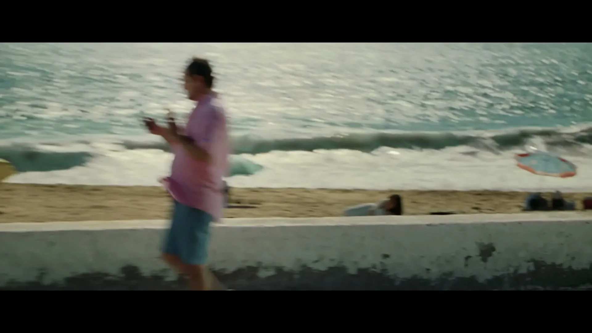 A man in a pink shirt and blue shorts walks along a beach wall, eating an ice cream and looking at his phone, as a security camera falls into the frame.