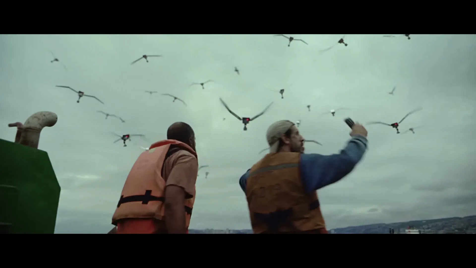 Two men in life vests on a boat look up at a flock of drones disguised as birds flying in a cloudy sky.