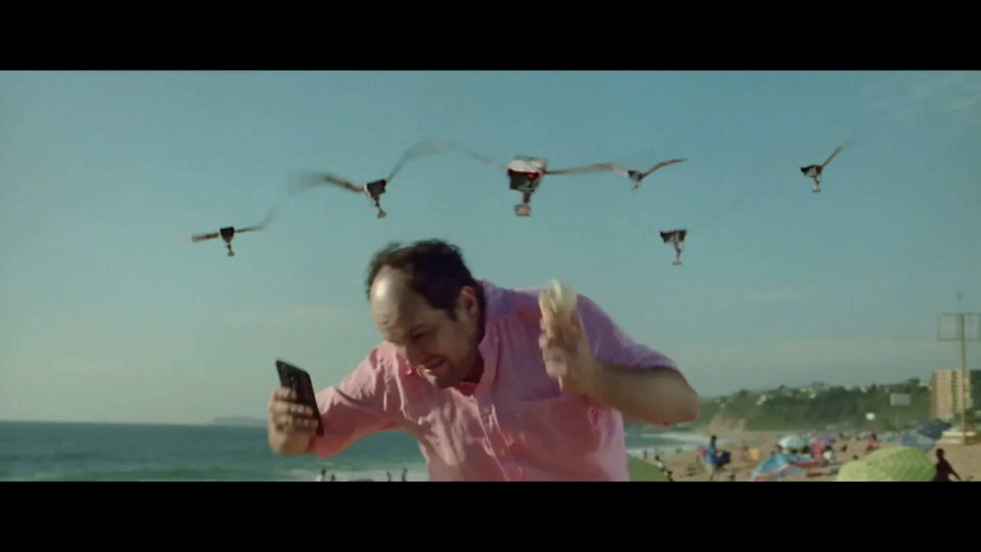 A man in a pink shirt runs on a beach, holding a phone and an ice cream, pursued by several bird-like drones.