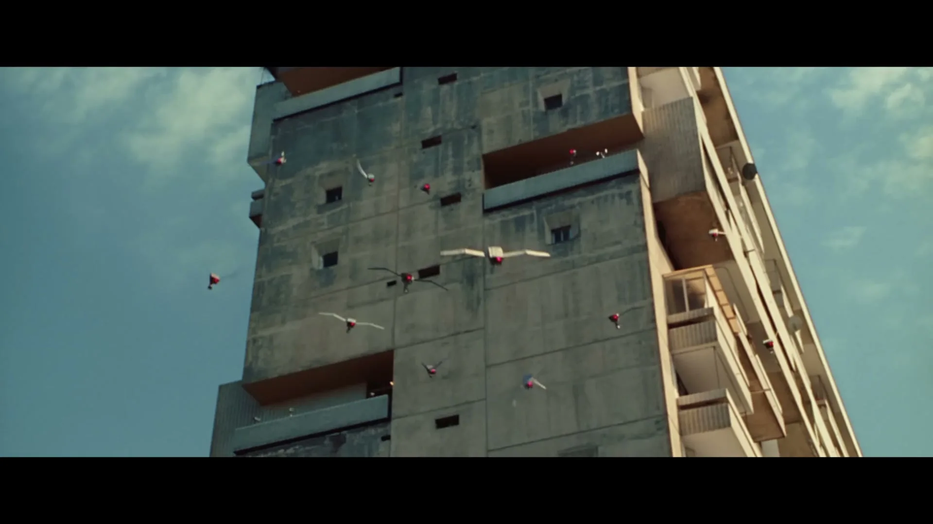 A flock of robotic birds with red lights on their heads fly around a brutalist concrete apartment building under a blue sky.