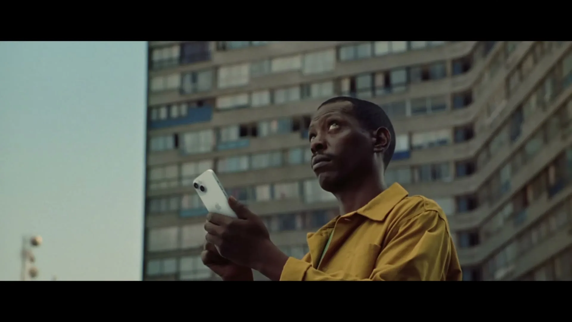 A Black man in his 30s, wearing a yellow shirt, looks up from his white iPhone against the backdrop of a large apartment building.