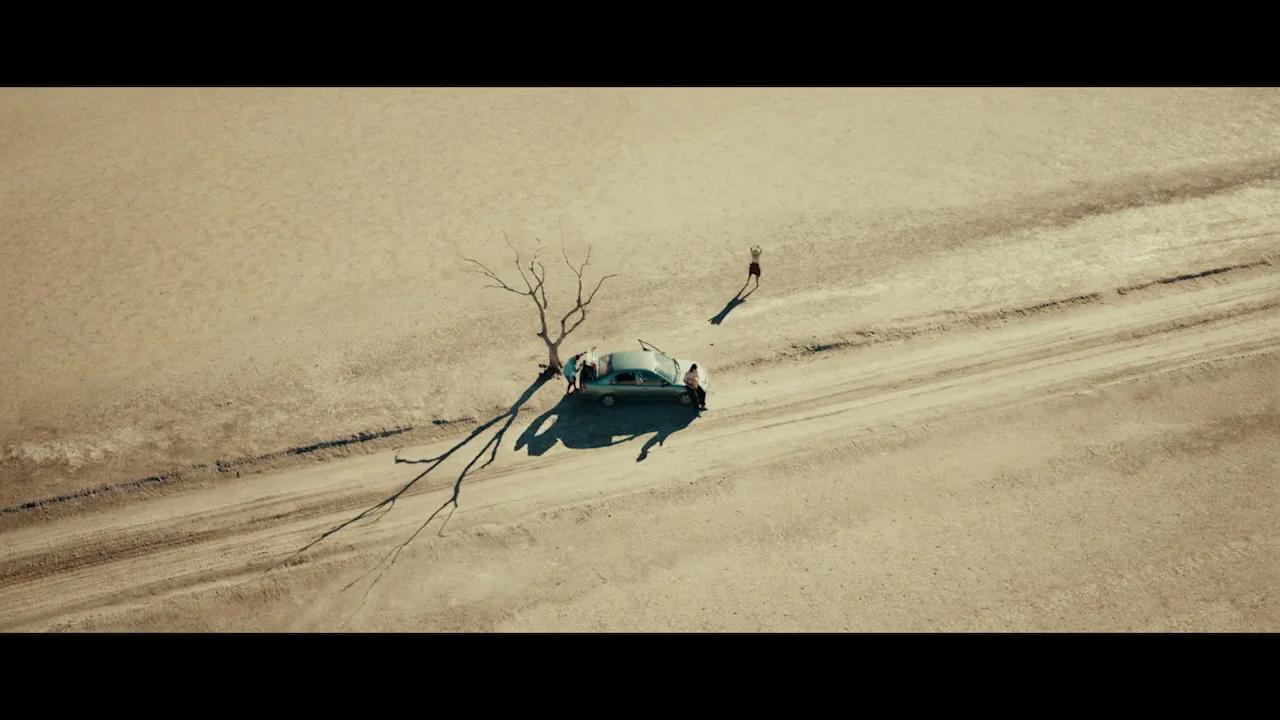 An aerial shot shows a broken-down car and a dead tree on a dirt road in a vast, empty desert landscape under a bright sky.