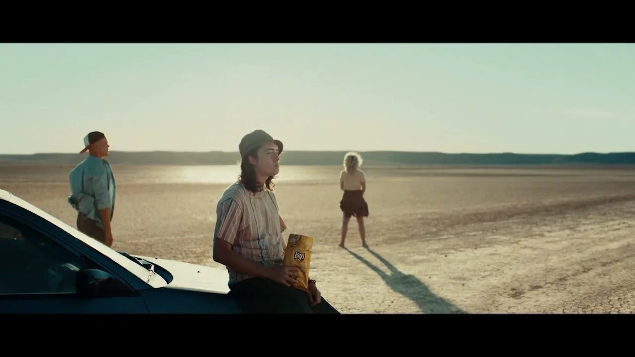 A young man sits on a car hood, eating chips, while a woman stands in the distance and another man walks towards them in a vast, dry landscape.
