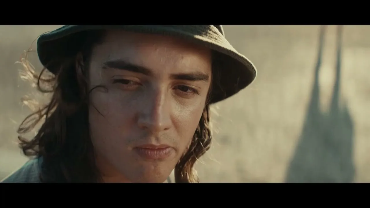 A young man with long hair and a bucket hat looks down with a slight frown, as wind blows his hair in a dusty, outdoor setting.