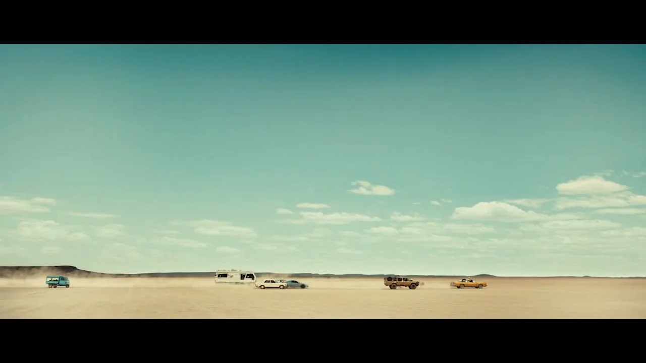 A wide shot shows several cars and an RV driving across a vast, dusty desert landscape under a bright blue sky with scattered clouds.