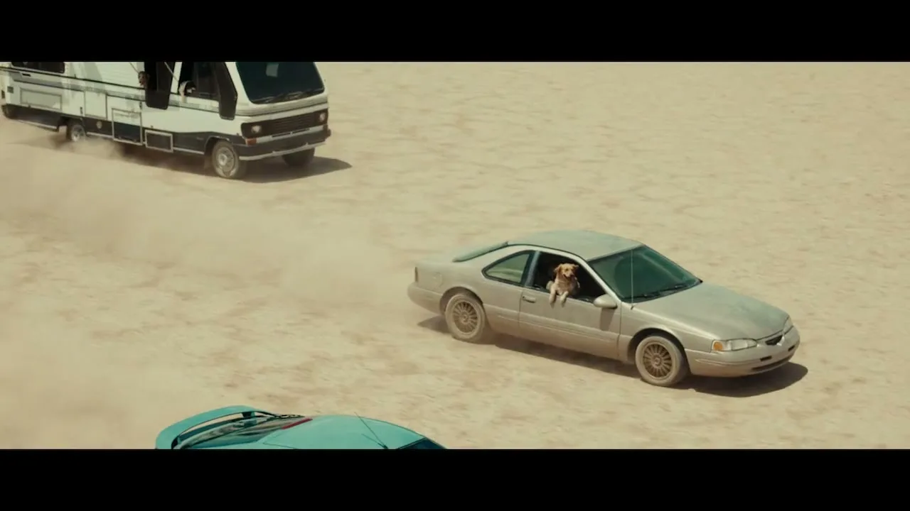 A golden retriever dog with its head out the window of a silver car, driving across a vast, dusty desert landscape with other vehicles.