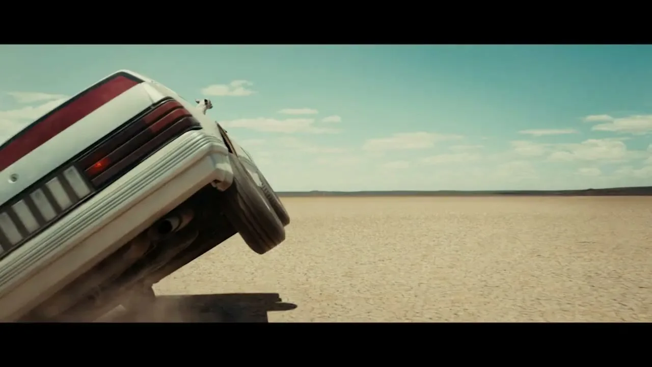 A white car drives on two wheels across a vast, dry desert landscape under a clear blue sky, kicking up dust.