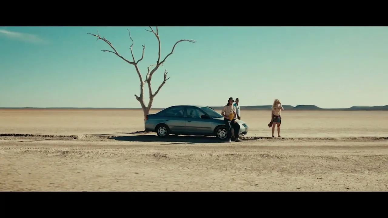 Three people stand by a car and a dead tree in a vast, arid desert landscape under a clear sky.