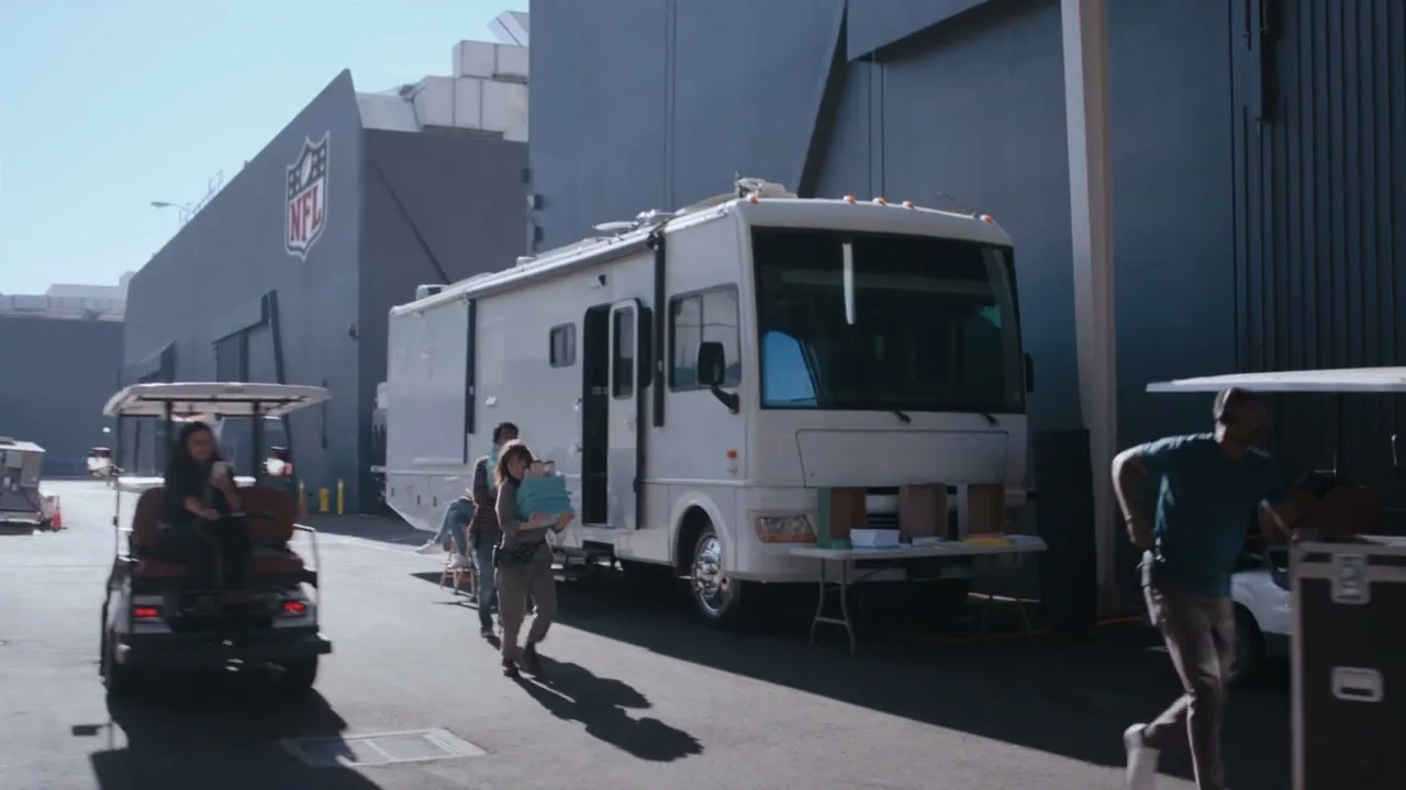 People arrive at a large building with an NFL logo, some on golf carts and others walking, carrying items, next to a large RV.