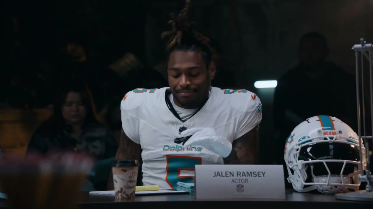 Jalen Ramsey, a male football player in a white Dolphins jersey, sits at a table with a football helmet and a name card in a dimly lit room.