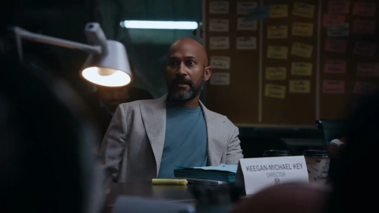 Keegan-Michael Key, a bald man with a beard, sits at a table in a dimly lit room, looking surprised.