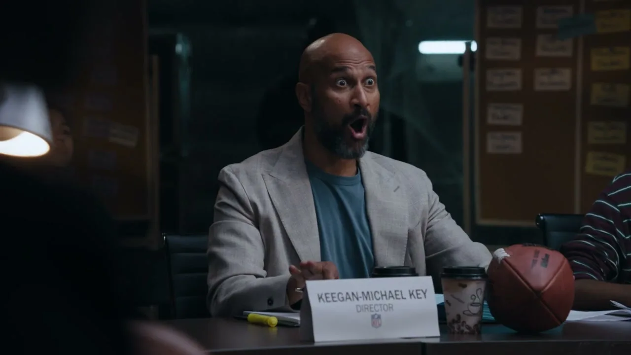 Keegan-Michael Key, a bald man with a beard, wearing a light grey suit jacket and blue t-shirt, sits at a table with a name card and a football, looking surprised in an office.