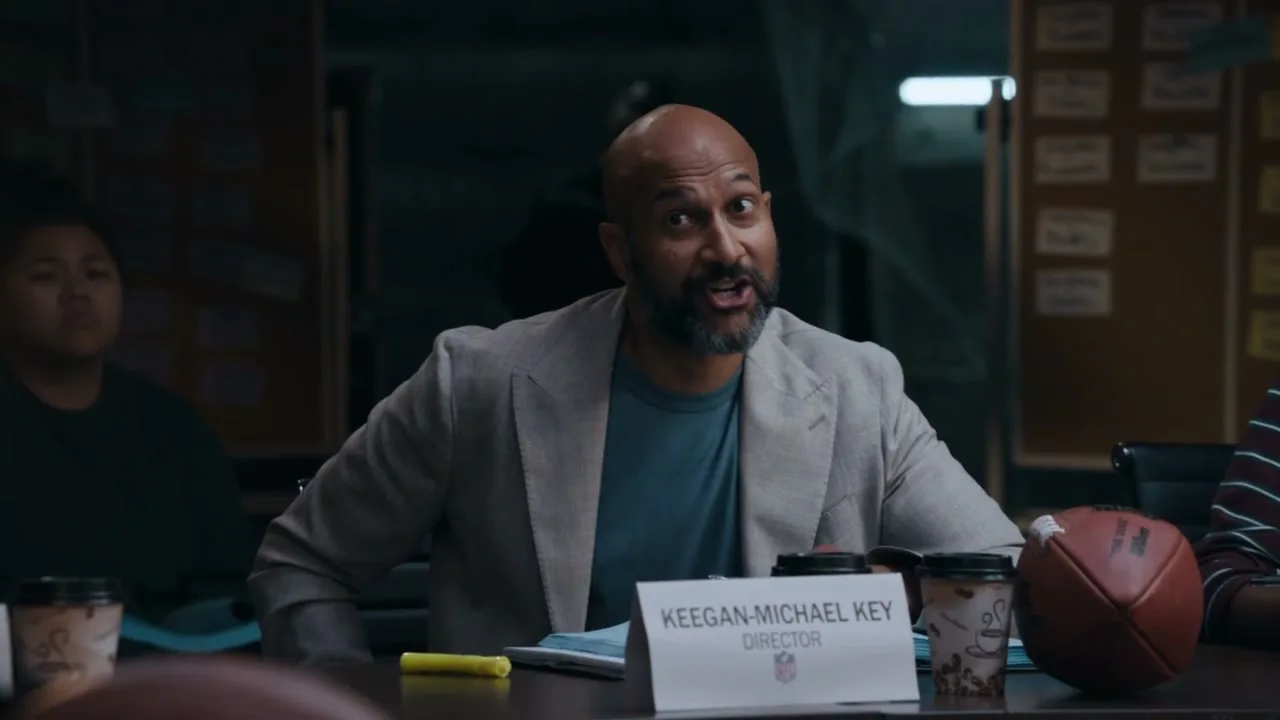 Keegan-Michael Key, a bald man with a beard, wearing a light grey suit jacket and blue t-shirt, sits at a table in a dark room, speaking.
