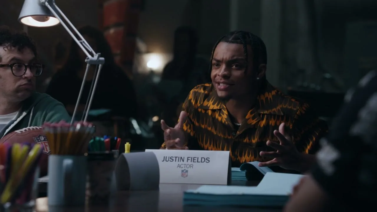 Justin Fields, an actor, sits at a table in a dimly lit room, wearing a patterned shirt and gesturing with his hands during a table read.