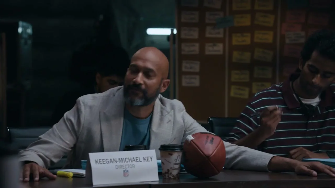 Keegan-Michael Key, a bald man with a beard, sits at a table with a football and coffee, talking during a meeting.