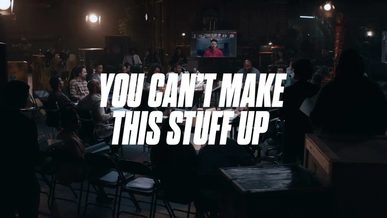 A wide shot of a dark soundstage with a large group of people sitting in chairs, with a video call on a screen in the background and white text overlayed.