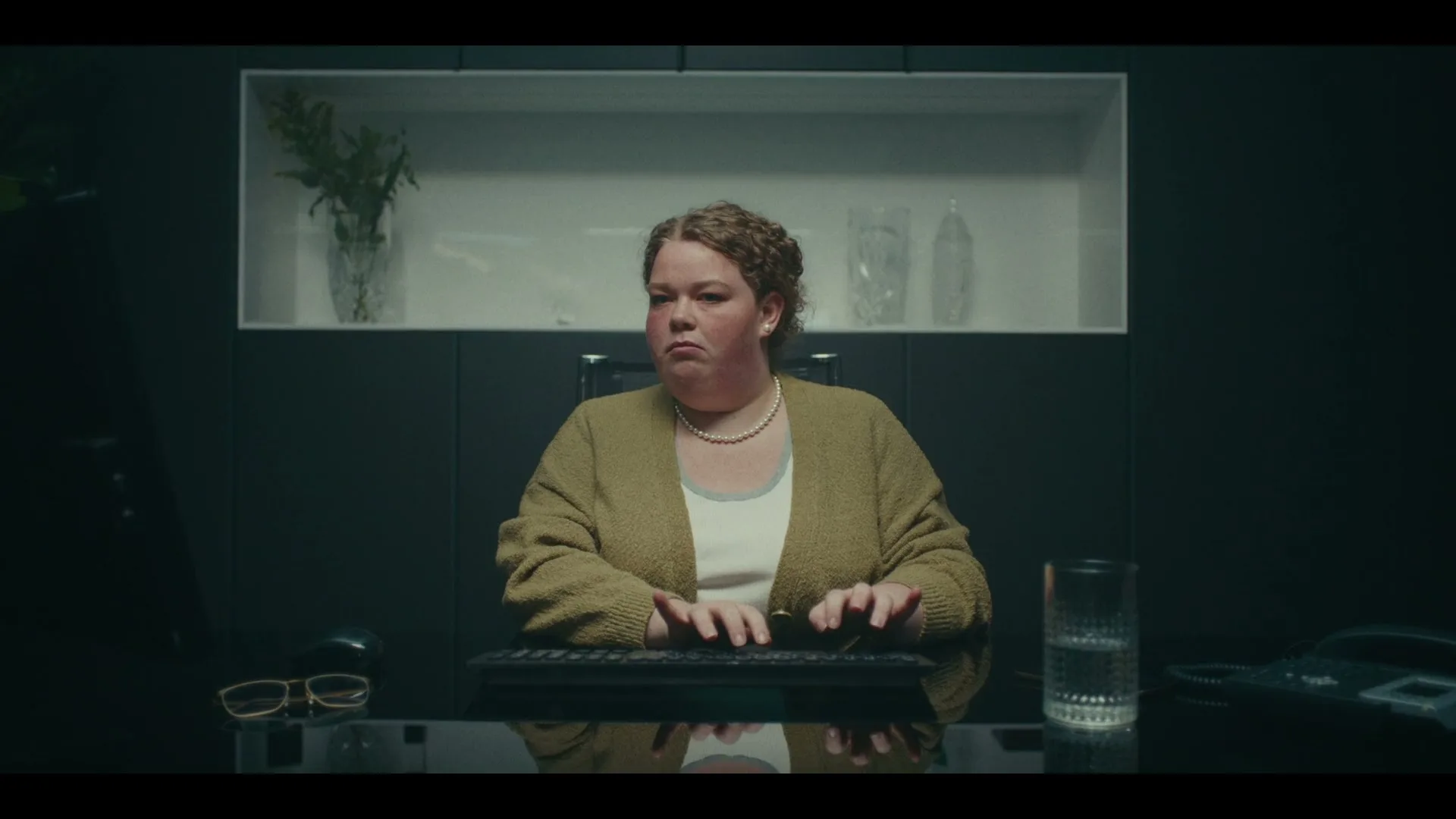A woman with curly hair and a pearl necklace sits at a desk, typing on a keyboard with a serious expression in an office.
