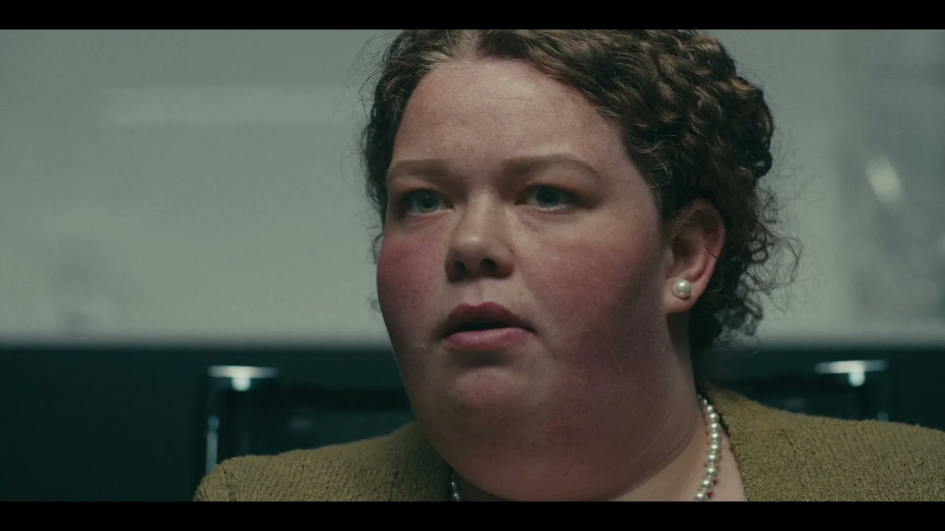 A woman with curly brown hair and a pearl necklace looks forward with a concerned expression in an interior setting.
