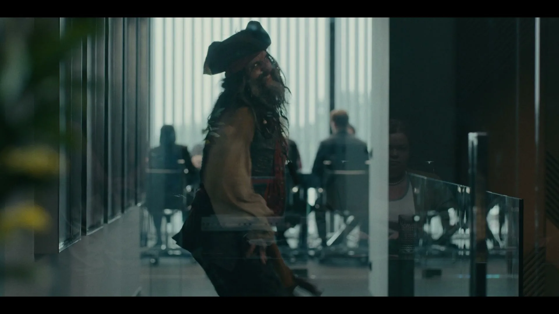 A man dressed as a pirate with a long beard and hat dances in an office building, reflected in a glass wall.
