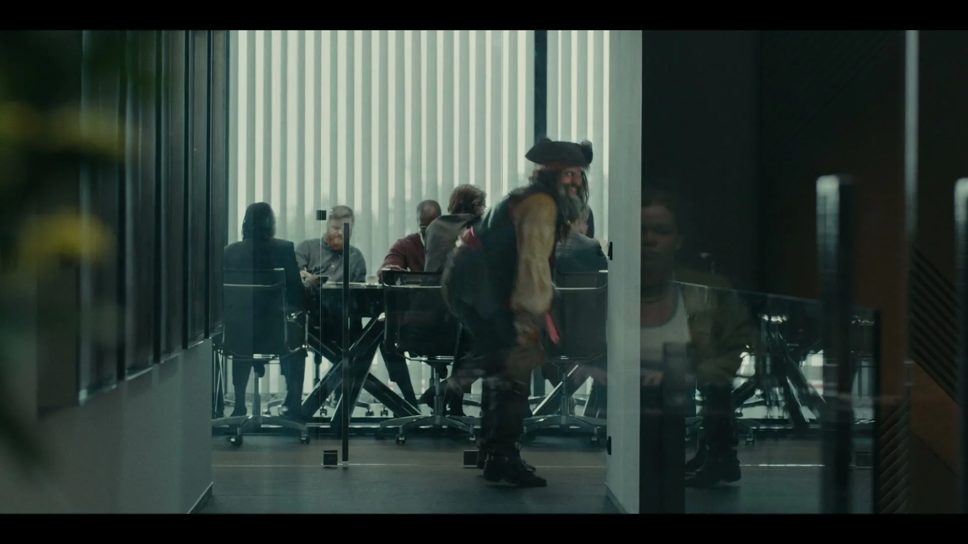A man dressed as a pirate walks past a glass-walled conference room where several people are seated at a table, in an office building.