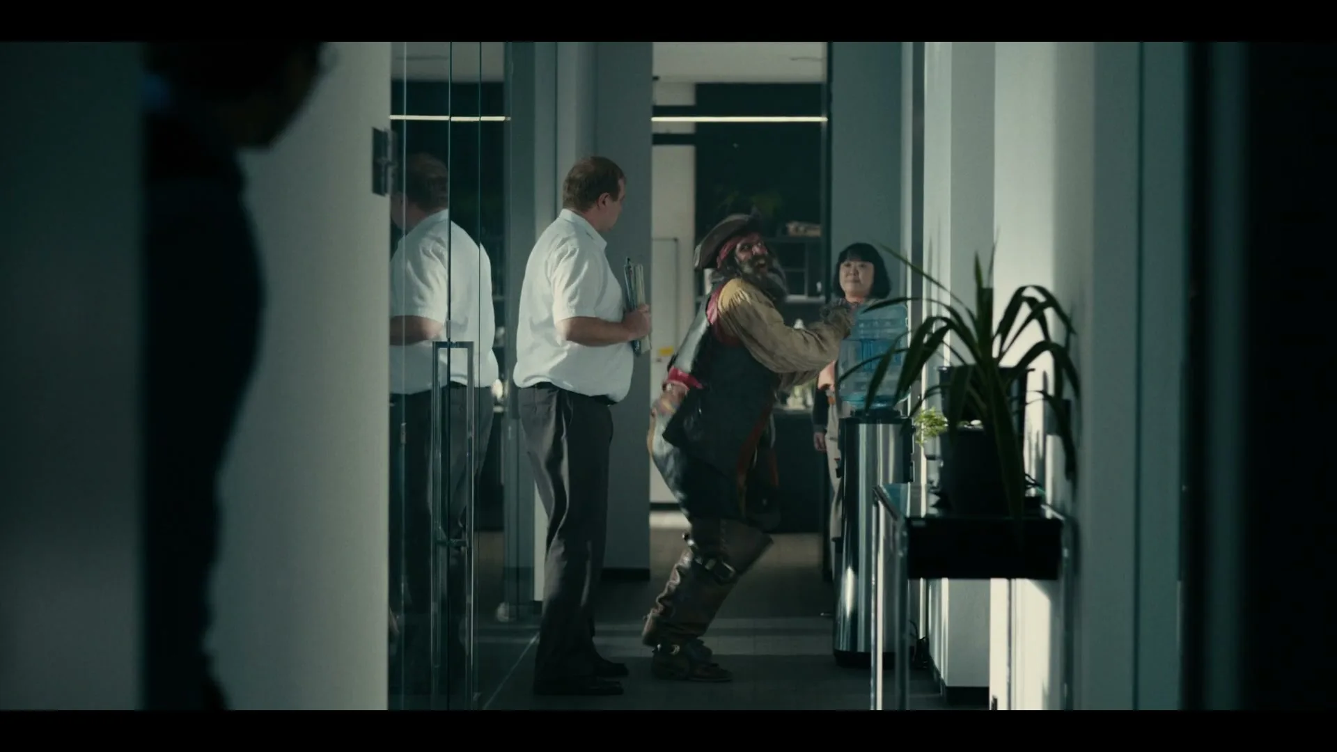 A pirate-costumed man tries to fill a water bottle at a water cooler in an office hallway, while two office workers watch him.