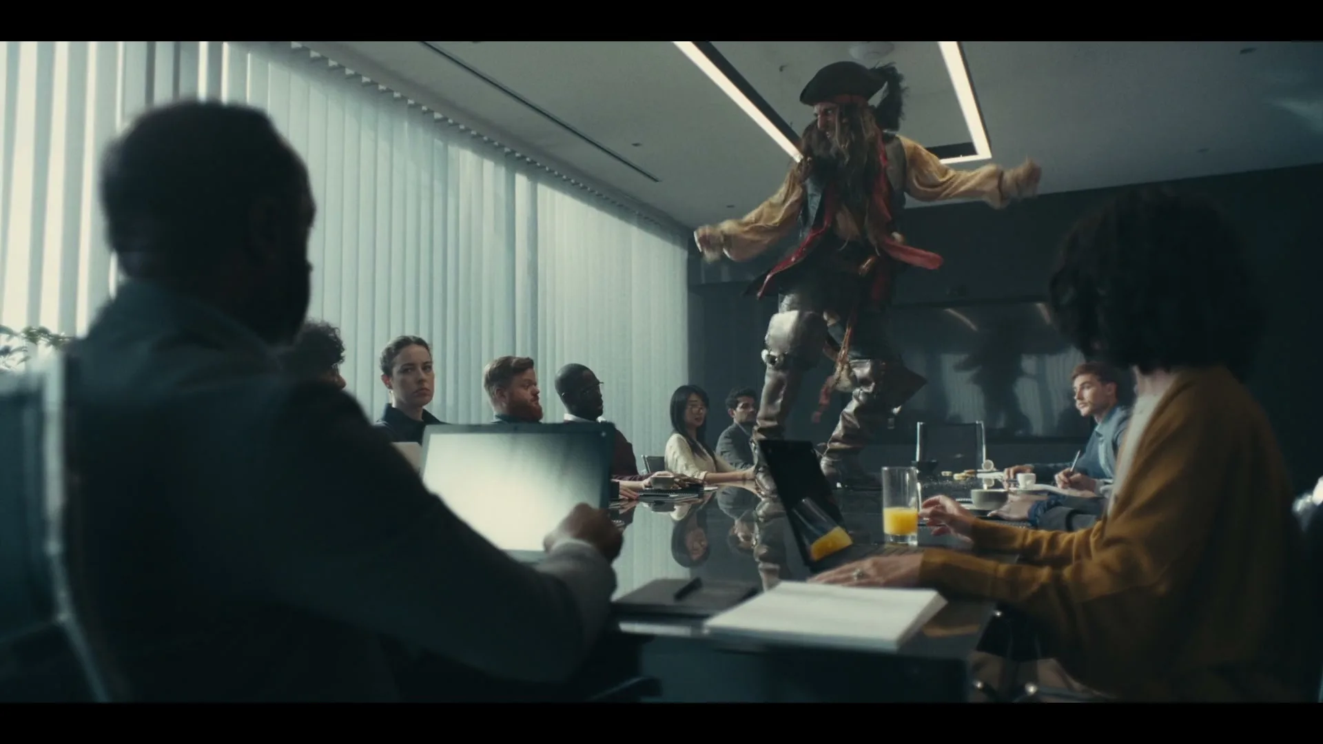A pirate-like figure in a tricorn hat and tattered clothes dances on a conference table while business people watch in an office.