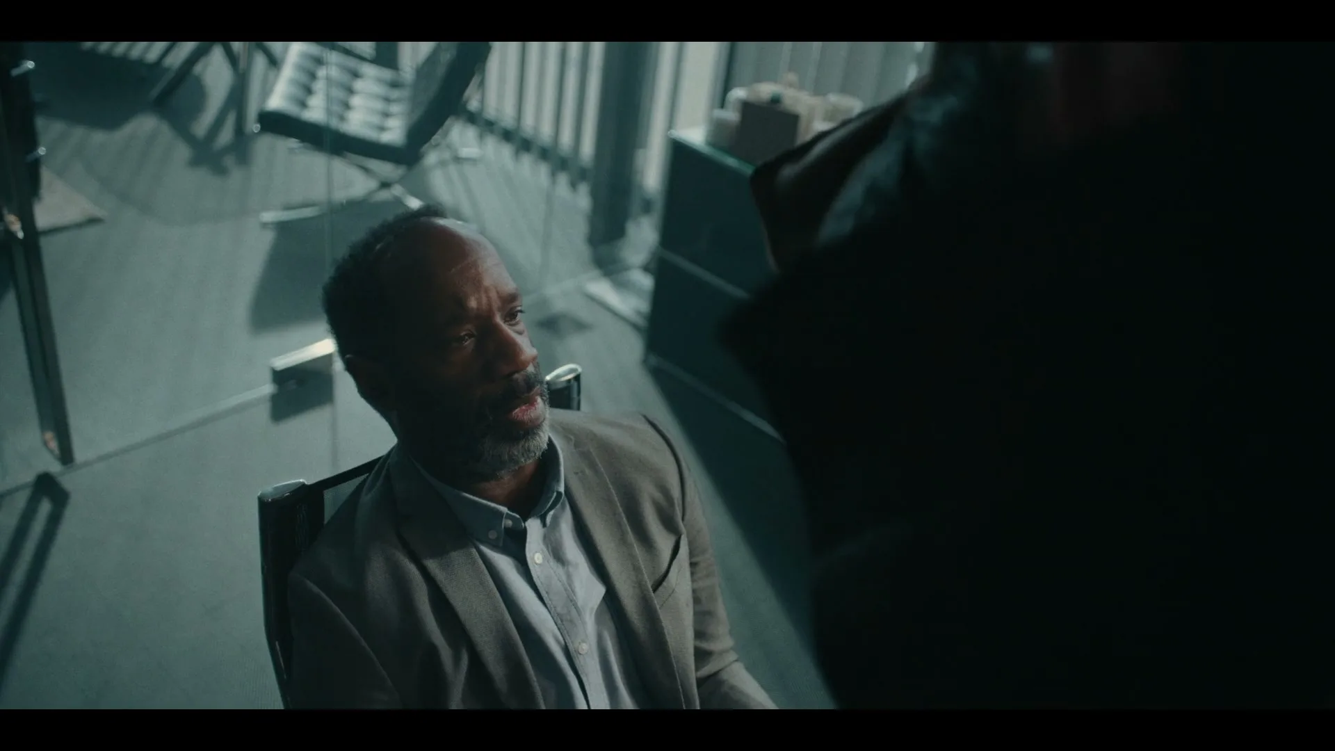 A man with a beard and a tan jacket sits in a chair, looking up at someone off-screen in a dimly lit room.