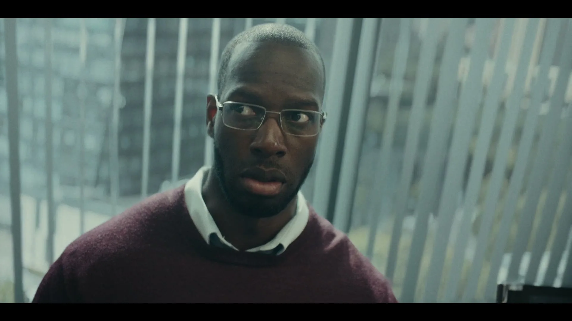 A Black man in glasses and a maroon sweater looks around nervously in an office building with vertical blinds.