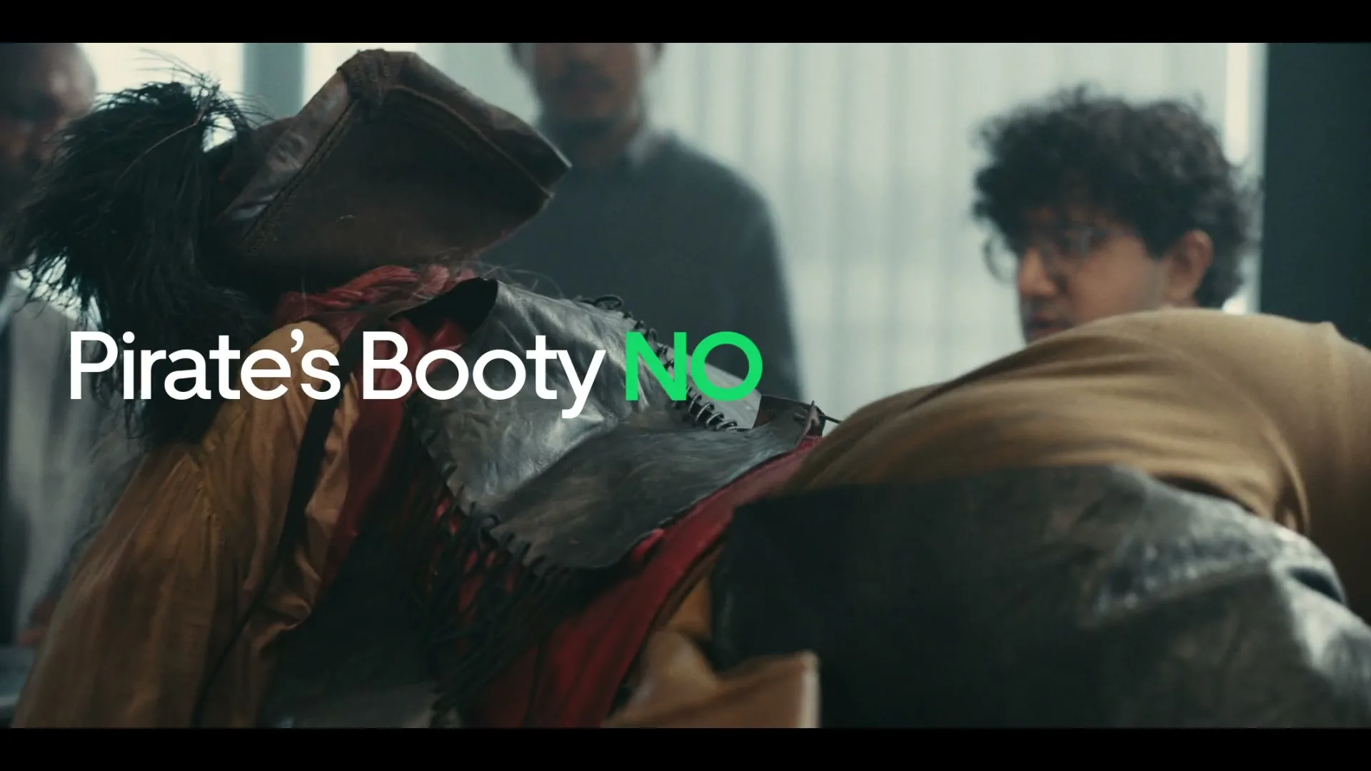 A group of people in an office watch a pirate character, dressed in a brown hat and red shirt, fall onto a table.