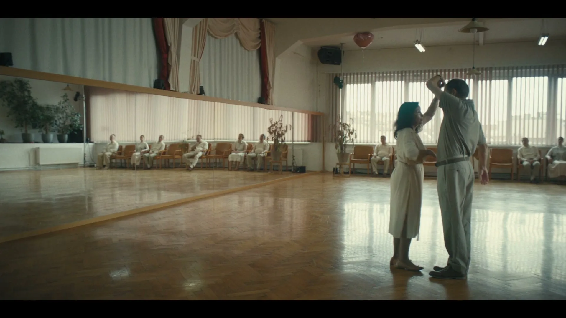 A long-haired older man sits at a table with a record player, watching a couple dance in a large, mirrored dance studio.