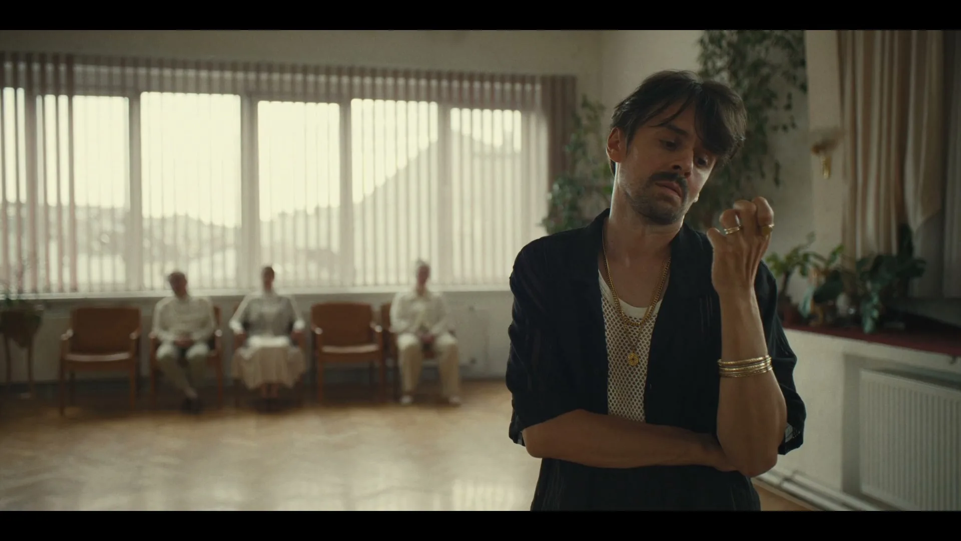 A man with a mustache and gold jewelry stands in a waiting room, looking down thoughtfully, while three elderly people sit in the background.