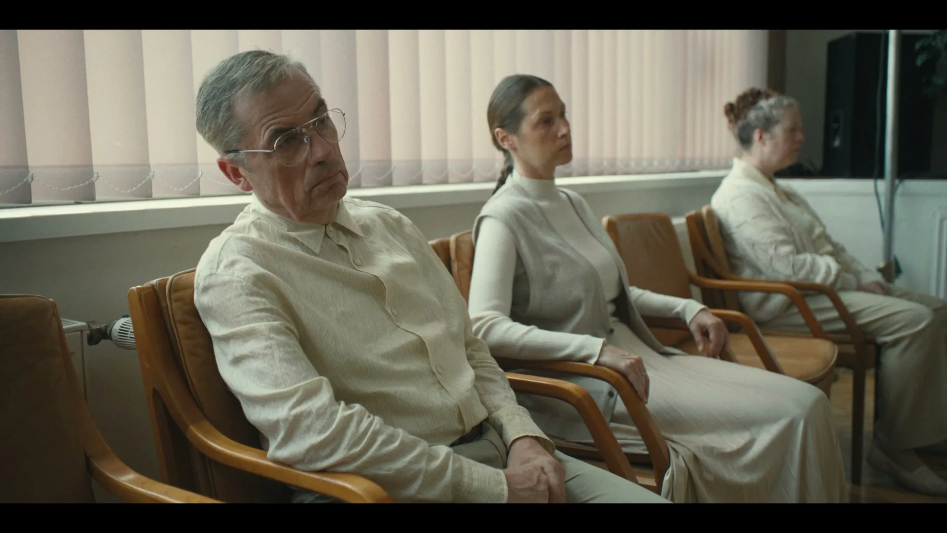 Three people, two women and one man, sit in a waiting room, all wearing light-colored clothing and looking bored or pensive.