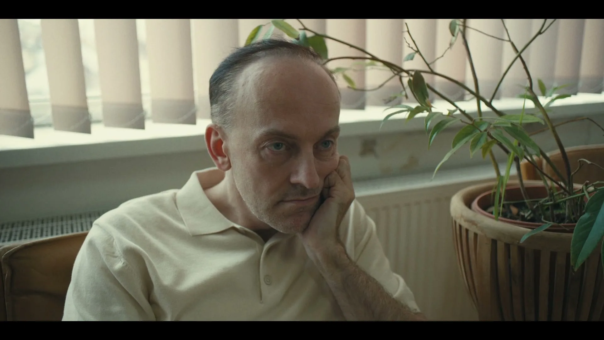 A middle-aged man with a receding hairline, wearing a light polo shirt, sits with his hand on his chin, looking bored or pensive indoors.