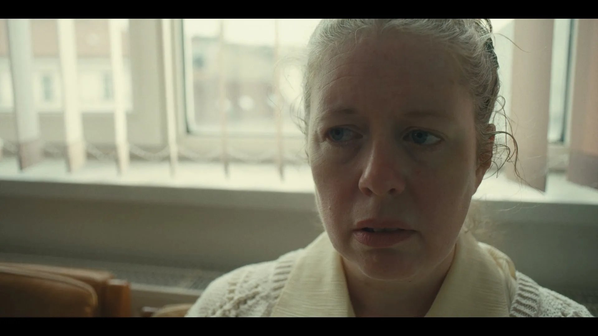 A middle-aged woman with blonde hair and a white sweater looks distressed, sitting indoors by a window with vertical blinds.