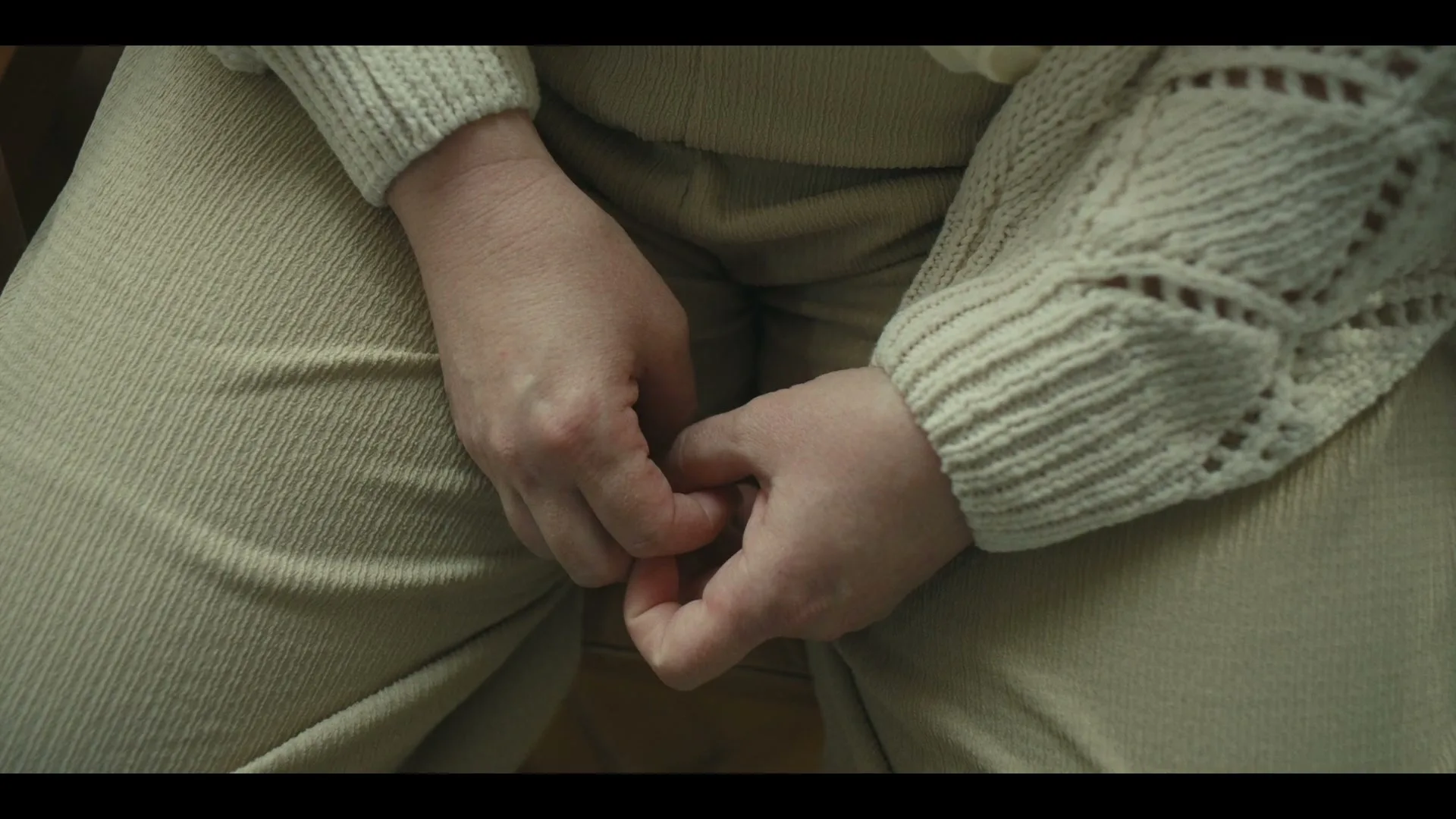 A person's hands are clasped together, resting on their lap, wearing a cream-colored knitted sweater and beige pants.
