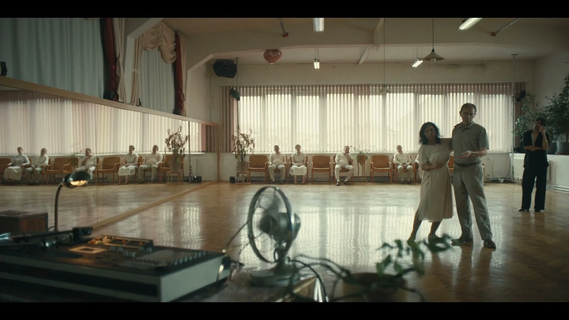 A man and a woman dance in a large, brightly lit room with a mirrored wall, while several people in white robes sit watching in the background.