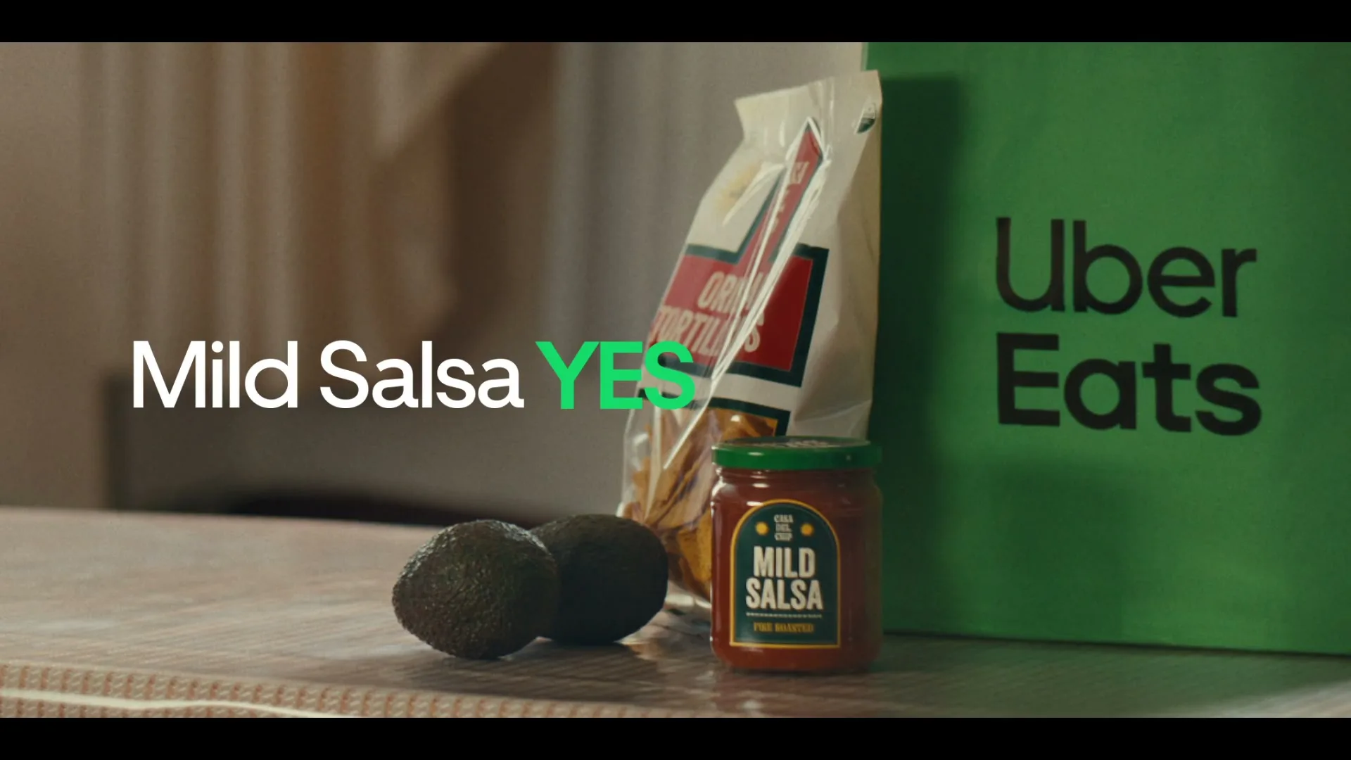 A jar of mild salsa, a bag of tortilla chips, and two avocados sit on a wooden surface next to a green Uber Eats bag.