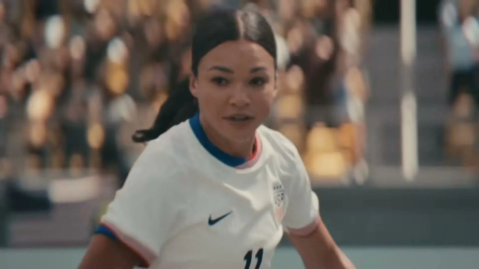 A female soccer player in a white jersey with a Nike logo and "USA" on it, runs on a field during a game.