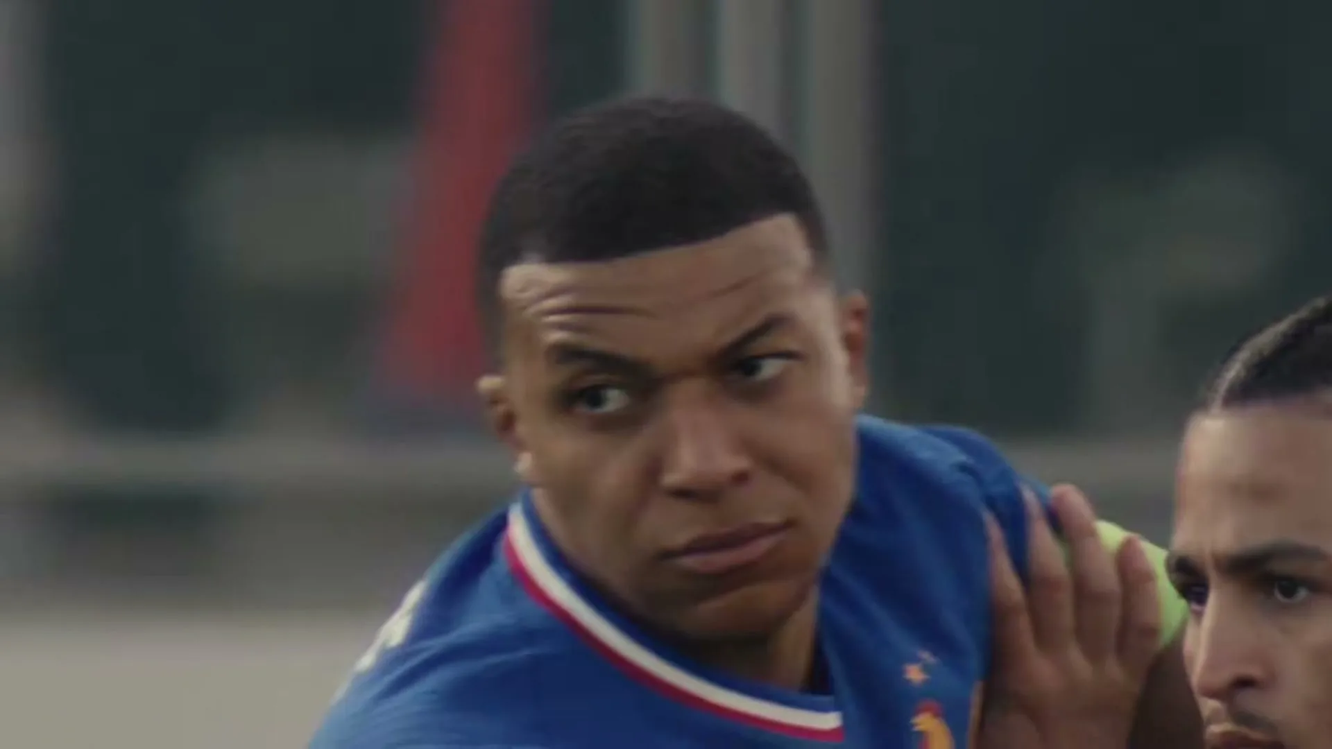 Kylian Mbappé, a male soccer player in a blue jersey, looks intently to his right while another player's arm is visible in the foreground.