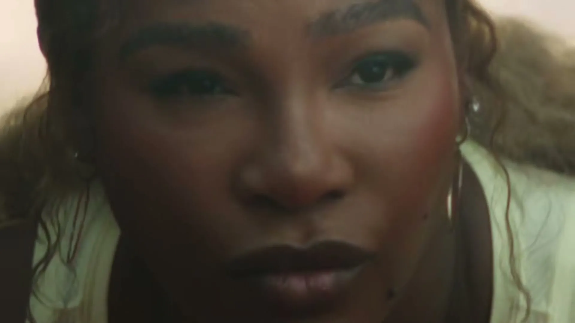 Serena Williams, a Black woman with long blonde hair, looks up at the camera with a serious expression.