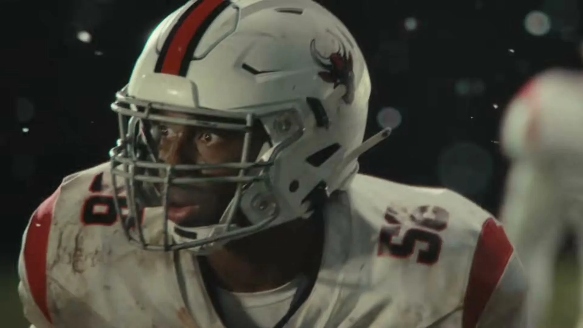 A young male football player in a white and red uniform with number 55, wearing a helmet, looks intensely forward on a dark field.