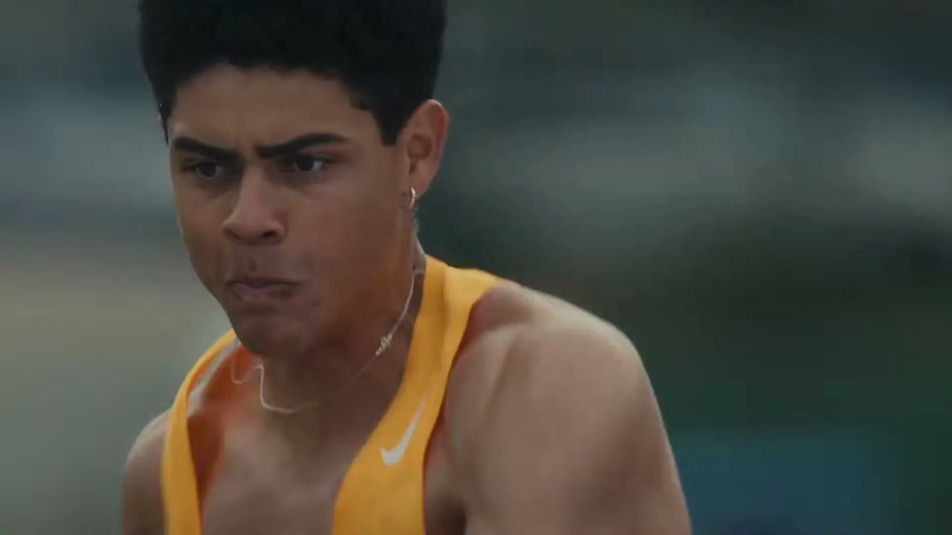 A determined male pole vaulter in a yellow tank top holds his pole, preparing for his jump at an outdoor track and field event.