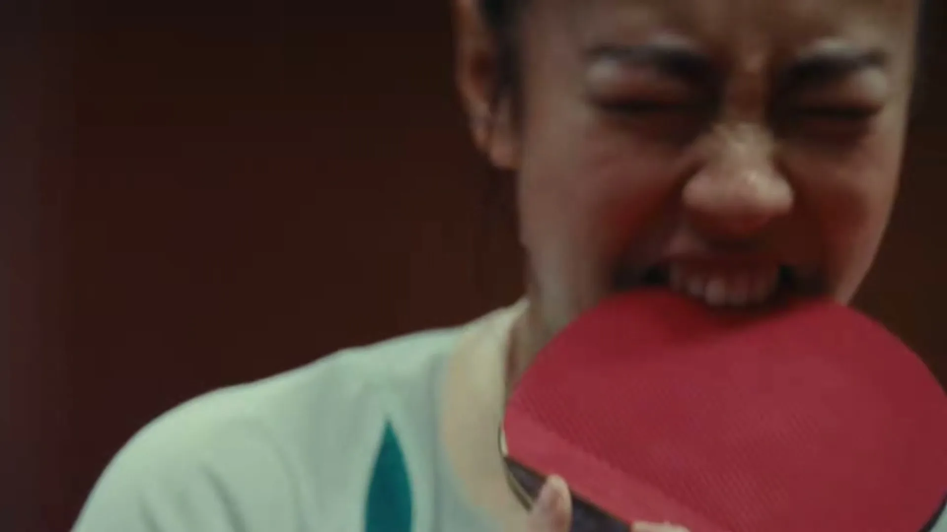 A female athlete in her 20s, wearing a white and green sports top, bites down on a red ping pong paddle with an intense expression, in an interior setting.
