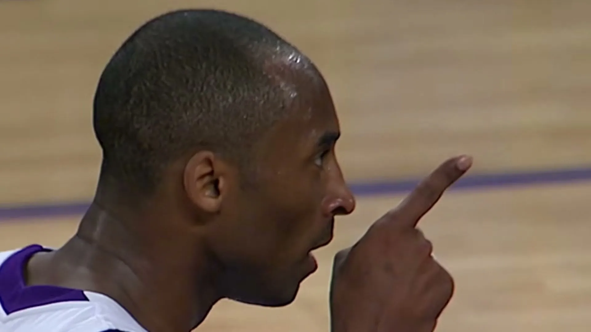 A male basketball player, sweating and wearing a white and purple jersey, points his finger while speaking on a basketball court.